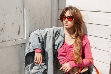 Moda mankeni, turuncu gözlüklü, şık, pembe bluzlu, kot ceketli güzel bir kadın plajdaki klasik beyaz ahşap binanın yanında duruyor.