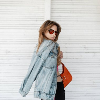 Havalı, genç, kızıl saçlı, şık güneş gözlüklü, modaya uygun kot ceketli, deri çantalı, sokakta beyaz ahşap bir duvarın yanında poz veren bir kız.