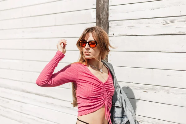 ファッショナブルなピンクの作物の上にファッションサングラスを持つスタイリッシュな若い女性の夏の女性のライフスタイルの肖像画と木製の白い壁の近くのビーチを歩くジーンズジャケット — ストック写真