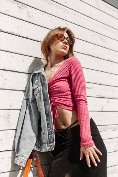 Beautiful sexy fashion woman with cool orange sunglasses in fashionable casual denim clothes with a pink crop top, jeans jacket, skirt and bag stands near a vintage white wooden wall on a sunny day