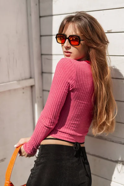 时尚漂亮的红头发女孩 戴着装饰过的眼镜 穿着粉色时髦运动衫 阳光灿烂 温暖的日子里 靠近古老的木制白墙 — 图库照片