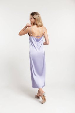 Modaya uygun, sarı saçlı, şık, mor, uzun, sırtı açık, ayakkabılı güzel bir kadın stüdyoda beyaz bir arka planda yürüyor.