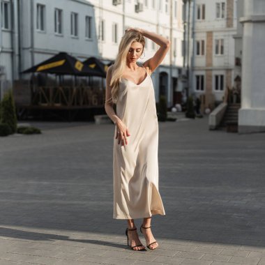 Şık, uzun, bej renkli, ayakkabılı zarif sarışın kadın gün batımında şehirde yürüyor.