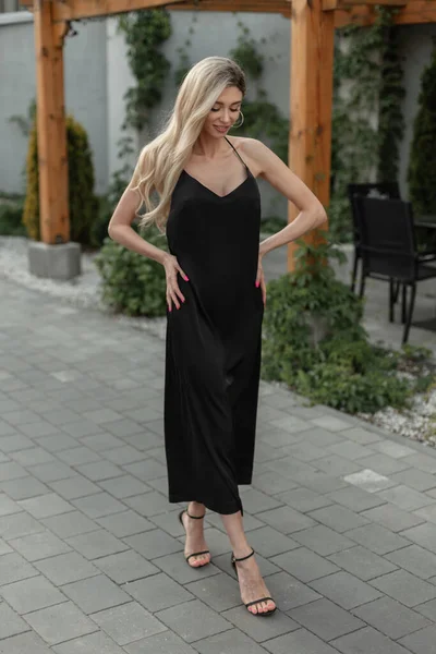 Fashion pretty elegant chic woman with blonde hair in stylish black long sexy strappy dress with shoes walks on the street