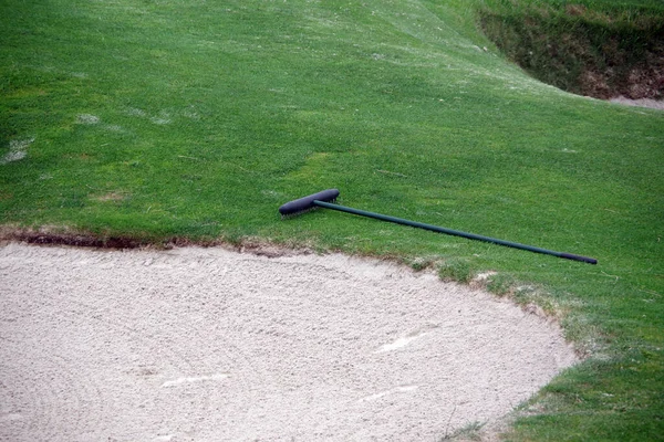 Partial view of a golf course sand trap bunker and a rake next to it on the grass