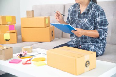 Asyalı bayan girişimci KME sahibi, müşteri siparişlerini kontrol ediyor ve ürünleri kutulara koyuyor. Çevrimiçi İş Fikirleri ve Serbest Çalışmalar