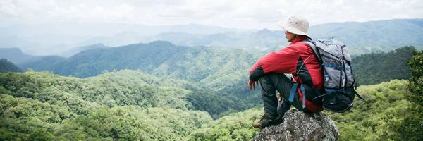 Young person hiking male on top rock, Backpack man looking at beautiful mountain valley at sunlight in summer, Landscape with sport man, high hills, forest, sky. Travel and tourism.