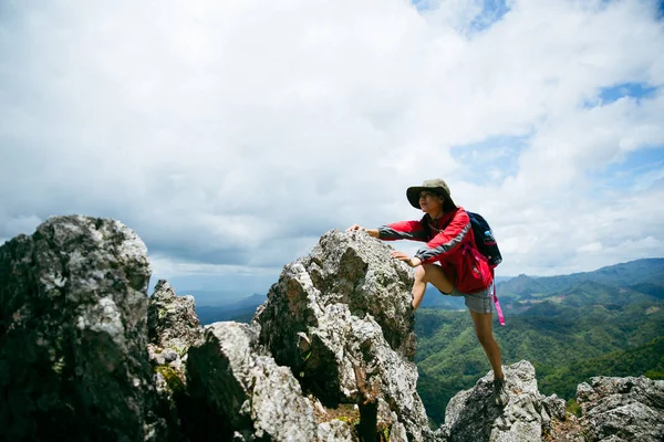 Young person hiking female climbing on top rock, Backpack woman looking at beautiful mountain valley at sunlight in summer, Landscape with sport girl, high hills, forest, sky. Travel and tourism.