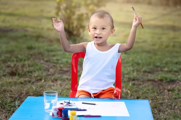 Kid Painting. Happy little cute child girl with dirty hands and fingers, using paintbrush painting on paper and her Tank top. Little girl painting water color in the garden on backyard.