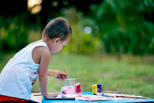 Çocuk boyama. Kirli elleri ve parmakları olan mutlu küçük kız çocuğu, boya fırçasını kağıt ve kolsuz bluzuyla boyuyor. Bahçede sulu boya yapan küçük bir kız..