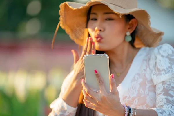 Focus on Phone. Happy Asian woman sit in the flowers farm outdoors and chitchat on online dating app in mobile phone. Women Online dating via video call  at Sunset. New normal valentine's day concept