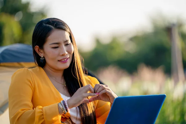 Happy beauty Asian woman in yellow dress sitting in the flowers farm outdoors and chitchat online thru laptop computer. Women Online dating via video call  at Sunset. New normal valentine\'s day concept