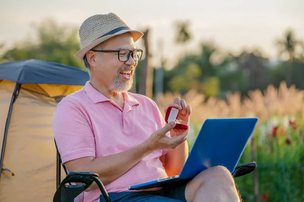 Mutluluk Adamı internetten kız arkadaşına dizüstü bilgisayarla sürpriz evlilik teklifi yapıyor. Gri sakallı emeklilik ya da Sunset 'teki yaşlı adam. Karantina Sevgililer Günü konsepti.