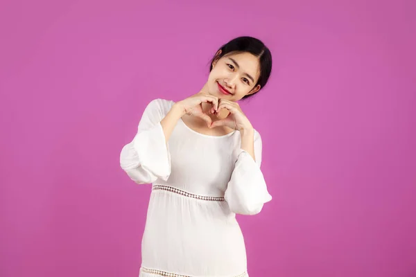 若いです可愛いですアジアの女性でホワイトドレスとともに手ハートサインオンザピンクスクリーン背景 — ストック写真