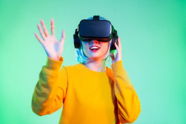 Metaverse konsept, sarı kazaklı mutlu Asyalı kadın VR gözlük takıyor ve neon yeşil arka planda oynuyor ve izliyor.