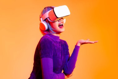 Metaverse konsept, mor eşofman altı giyen Asyalı genç kadın siber punk saç stili beyaz VR kulaklık takıyor, portakal rengi arka planda dokunmatik izleme oyununu sunuyor..