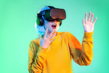 Metaverse konsept, sarı kazaklı genç Asyalı kadın VR kulaklık takıyor ve izole edilmiş neon yeşil arka planda bir şeye dokunuyor..