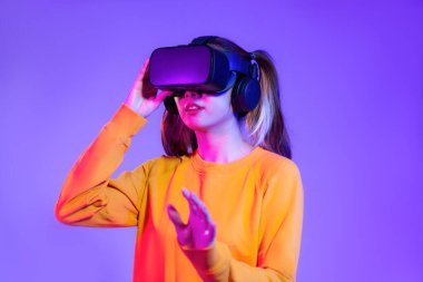 Siyah VR kulaklık takan turuncu kazaklı genç Asyalı kadın mor arka planda oynarken ve el ele tutuşurken izliyor. Gelecekteki kız yaşam tarzları hakkında metaverse konsept