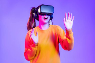 Turuncu kazaklı, siyah VR kulaklık takan mutlu genç bir kadın mor arka planda oynarken ve el ele tutuşurken izliyor. Gelecekteki kız yaşam tarzının değişken konsepti.