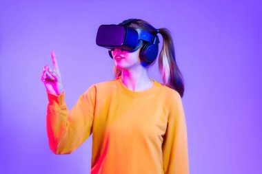 Turuncu kazaklı, siyah VR gözlüklü genç Asyalı kadın mor arka planda oynarken ve el ele tutuşurken izliyor. Gelecekteki kız yaşam tarzının değişken konsepti.