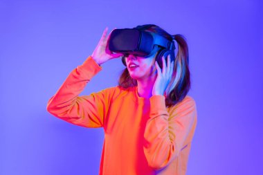 Siyah VR kulaklık takan turuncu kazaklı genç Asyalı kadın mor arka planda oynarken ve el ele tutuşurken izliyor. Gelecekteki kız yaşam tarzının değişken konsepti.