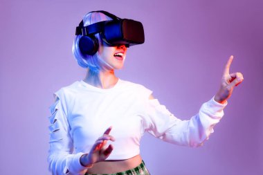 Siyah VR kulaklık takan beyaz tişörtlü genç Asyalı kadın mor arka planda oynarken ve el ele tutuşurken izliyor. Metaverse konsepti.
