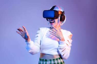 Siyah VR kulaklık takan beyaz tişörtlü genç Asyalı kadın mor arka planda oynarken ve el ele tutuşurken izliyor. Metaverse konsepti.