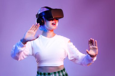 Siyah VR kulaklık takan beyaz tişörtlü genç Asyalı kadın mor arka planda oynarken ve el ele tutuşurken izliyor. Metaverse konsepti.