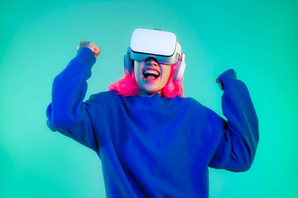 Young asian punk girl pink color hair style in blue sweatshirt wearing white vr headset watching playing online game on green screen background.
