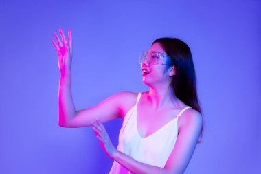 Young asian woman in white tank top wearing vr glasses posing touching on the bllue color background.