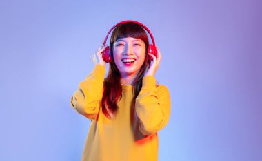 Sarı kazaklı genç Asyalı kadın kırmızı kulaklık takıyor gülümsüyor ve mor ekranda müzik dinliyor..