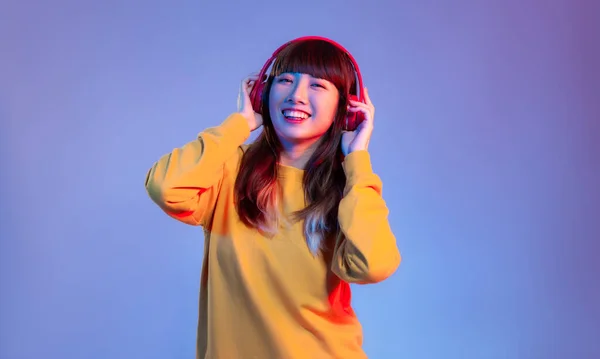Young asian woman in yellow sweatshirt wearing red headphones listen to music on the purple screen background.