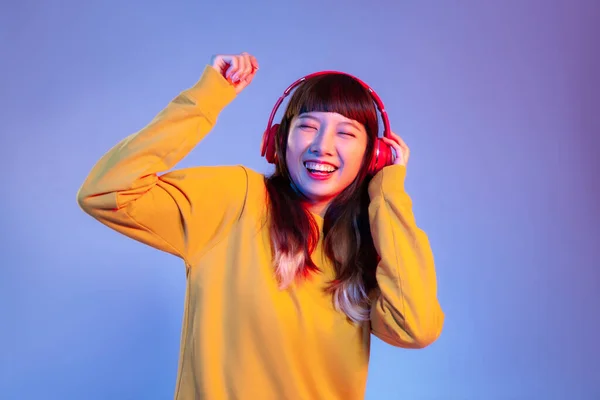 Young asian woman in yellow sweatshirt wearing red headphones listen to music and dancing on the purple screen background.