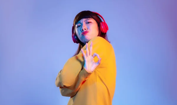 Young asian woman in yellow sweatshirt wearing red headphones listen to music on the blue screen background.