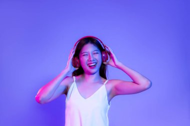 Young asian woman in white tank top wearing red headphone listen to music smiling on the purple neon light background.	