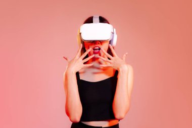 Metaverse konsept, siyah kolsuz bluzlu genç Asyalı kadın VR kulaklık takıyor kırmızı pastel ekranda heyecan verici poz veriyor..