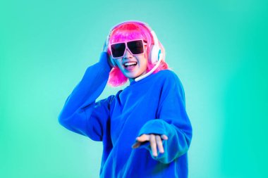 Mavi kazaklı genç Asyalı kadın pembe kısa saçlı punk tarzı güneş gözlüğü takıyor ve kulaklık takıyor yeşil ekranda dans ediyor..