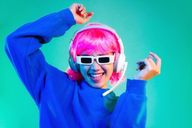 happy asian woman in blue sweatshirt pink short hair punk style wearing sunglasses and headphones dancing on the green background.	