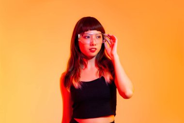 Young asian girl in black tank top wearing vr clear glasses posing on yellow screen background.
