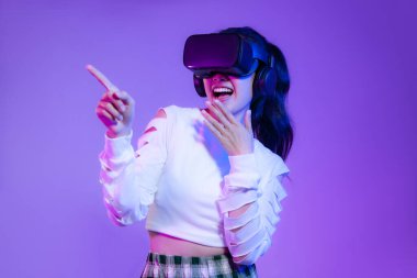 Young asian girl in wearing vr headset watching playing and finger pointing on the purple screen background.	
