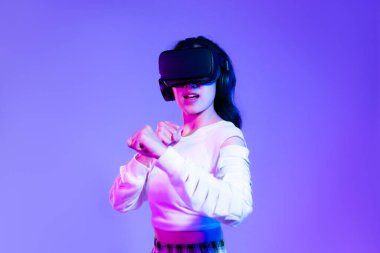 Young asian woman in white fashion sweatshirt wearing black vr headset posing watching and playing on the purple neon color screen background. Metaverse concept.