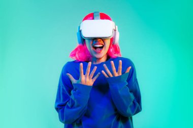 Genç Asyalı kız mavi kazaklı pembe saç stili beyaz VR kulaklık takıyor yeşil ekranda oynuyor ve sürpriz yapıyor. Cyborg ve Metaverse konsepti.
