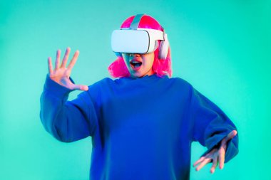 Young asian girl pink color hairstyle in blue sweatshirt wearing white vr headset watching playing and touching on the green screen background. Cyborg and Metaverse concept.