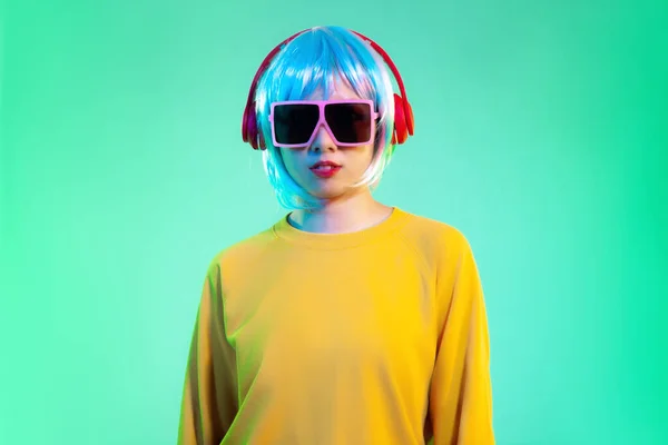Young asian girl in yellow sweatshirt blue color shot hair style wearing red headphone and pink sunglasses listen to music posing standing on the green screen background.