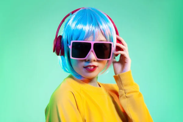 Young asian girl in yellow sweatshirt blue color shot hair style wearing red headphone and pink sunglasses listen to music posing looking on the green screen background.