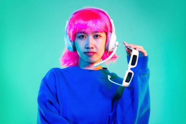 Young asian woman in blue sweatshirt pink short hair punk style wearing headphones holding sunglasses posing on the green screen background.