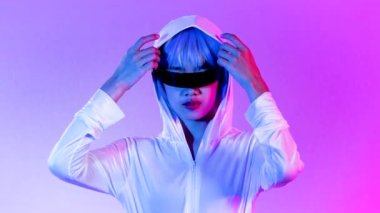 Metaverse gamer girl concept, young asian woman in white dress wearing 3d glasses posing on the neon purple color background. 4K video