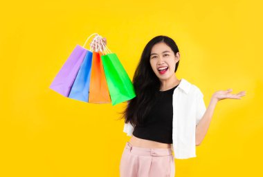 Young asian woman long hair style in black and white costume carrying the colorful paper shopping bags on yellow background with hand pointing for copy space.