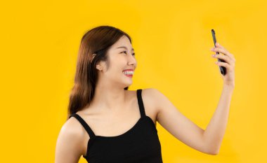 Young asian woman long hair in black tank top using cellphone chatting and selfie on yellow background.