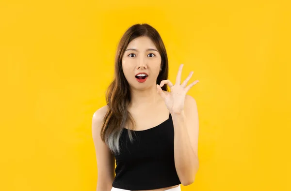 Young asian woman in black tank top posing hand okay sign on yellow background.
